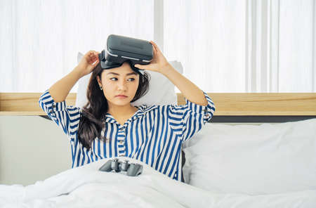 Asia woman wearing VR glasses and sitting in bed in the bedroom. Holding joy stick and playing game...