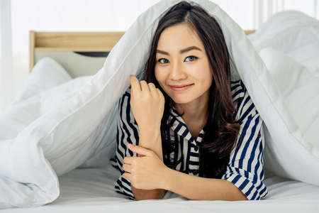 Young asian woman waking up at morning, Smiling woman resting under a duvet in her bedroom. Close Up portrait of a beautiful young woman and under the blanket. happy good morningの写真素材