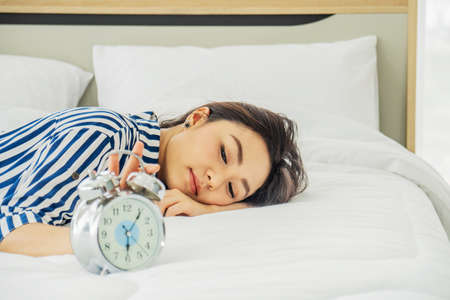 Beautiful and healthy young women waking up and her hand touching alarm clock on the bed, Lazy time and Holiday concept.の写真素材