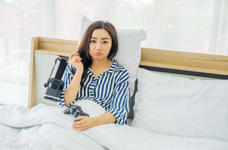 Bored asia woman Holding VR glasses and joy stick after playing game by using VR glasses sitting in bed in the bedroom, quarantine due coronavirus pandemic. stay at home.の写真素材