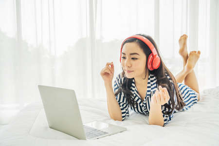 Beautiful young woman relaxing and listening the music while using laptop computer on the bed in bed room with copy space. Beauty asian girl gets happy, relaxing. She enjoying lifeの写真素材