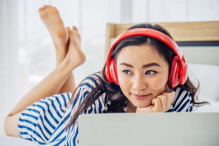 Beautiful young woman relaxing and listening the music while using laptop computer on the bed in bed room with copy space. Beauty asian girl gets happy, relaxing. She enjoying lifeの写真素材