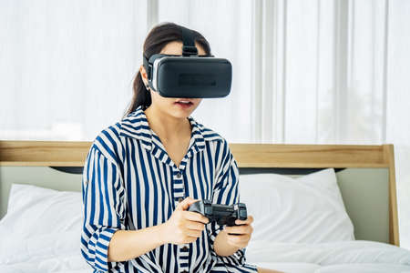 Asia woman wearing VR glasses and sitting in bed in the bedroom. Holding joy stick and playing game by using VR glasses, quarantine due coronavirus pandemic. stay at home.の写真素材
