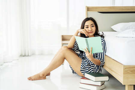Corona Virus, Covid-19, Coronavirus. Stay at home.Quarantine. 
Beautiful asian woman wearing sleepwear reading a book and sitting near bed in morning. Resting and Relaxation timeの写真素材