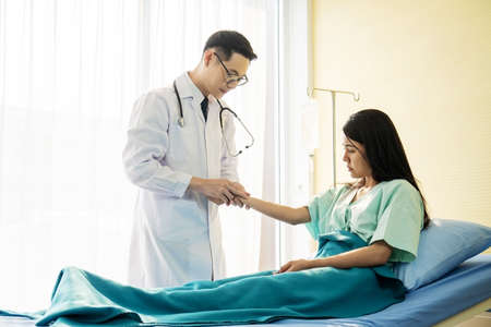Asian male doctor wearing stethoscope and check the pulse of a female patient sitting on a hospital bedの写真素材