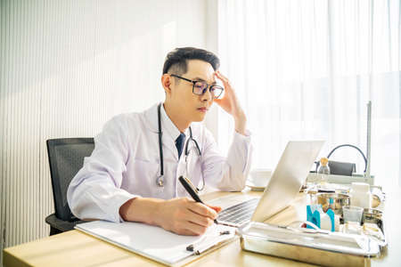Troubled asian man doctor working on computer at desk in hospital clinic, worried, thinking hard, healthcare concept for Covid19.の写真素材