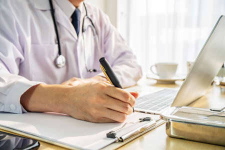 Side view cropped shot of male doctor in white uniform with stethoscope on neck at paperwork with notebook and laptop in the hospital.の写真素材