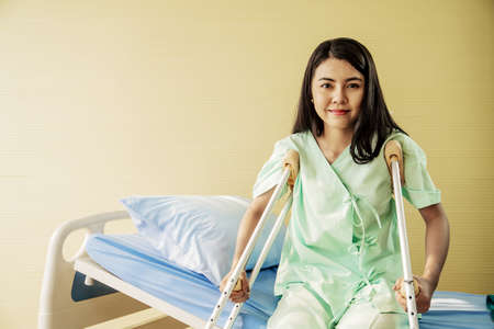Portrait of Happy Young female patient while sitting in bed with crutches in a hospital room. Health care and medical insurance concept.の写真素材