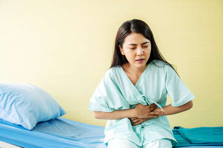 Woman patient  suffering from stomach ache while sitting on bed at hospital, abdominal pain, food poisoningの写真素材