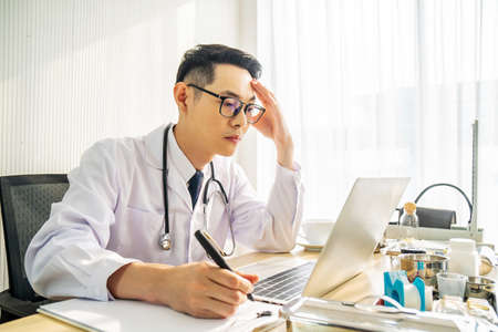 Troubled asian man doctor working on computer at desk in hospital clinic, worried, thinking hard, healthcare concept for Covid19.の写真素材