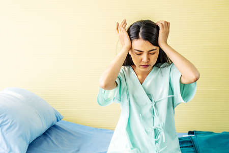 Asian young woman patients suffering headache with hands on forehead. medical and healthcare concept. sick frowning female sitting on bed in hospital room.の写真素材