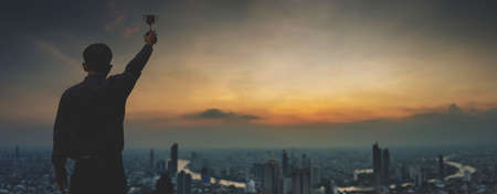 Success business concept, Silhouettes of Proud businessman holding up a golden trophy outdoors in sunset sky in front of the city skyline, corporate people individuals successful.の写真素材