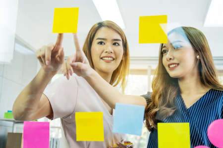 Two young business woman pointing sticky note papers on the glass thinking new idea with colleagueの写真素材