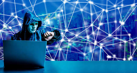 Anonymous computer hacker in white mask and hoodie. Obscured dark face using laptop computer for cyber attack and checking time  on modern city background, Data thief, internet attack,  darknet and cyber security concept.の写真素材