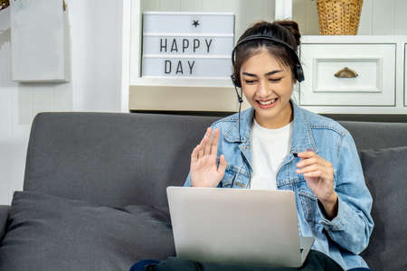 Asian young business woman is sitting on the sofa in her own house is wearing headphones video call conference with laptop computer raise hand saying hi team at home office, work from homeの写真素材