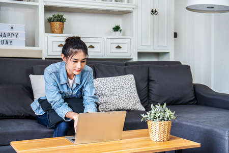 Asian teenage woman sitting on a sofa in the house is enjoying the work by using the computer laptop to make money from online business, work from homeの写真素材
