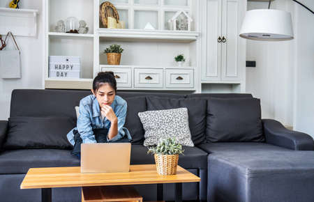 Asian teenage woman sitting on a sofa in the house is enjoying the work by using the computer laptop to make money from online business, work from homeの写真素材