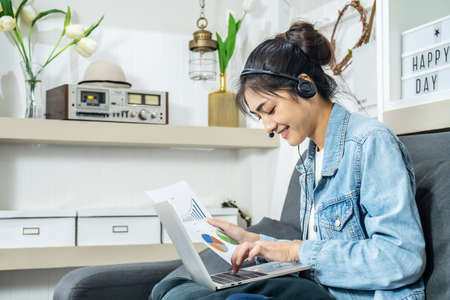 Asian young business woman is sitting on the sofa in her own house is wearing headphones video call conference with laptop computer with pile of graph and chart paper at home office, work from homeの写真素材