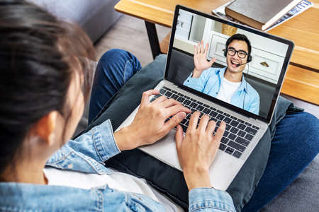 Back view of Asian business woman sitting on the sofa in her own house talking to a colleague about a video conference plan, Business team using laptop for a online meeting in video callの写真素材