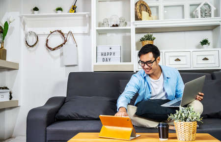 Smart Asian businessman in casual clothing sitting on a sofa in the house is working on computer laptop and tablet , SME start up concept, work from homeの写真素材