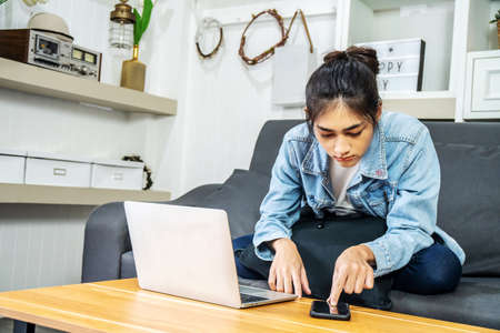Young Asian women sit on the sofa to work at home. Looking for information on laptops and mobile phones to get the job done, work from home conceptの写真素材