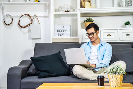 Smart Asian businessman in casual clothing sitting on a sofa in the house is enjoying the work by using the computer laptop to make money from online business, work from homeの写真素材