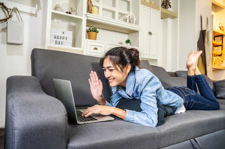 Asian woman is resting on the sofa in her own house, chatting on a video call with a close female friend on the computer laptop screen, Working from homeの写真素材
