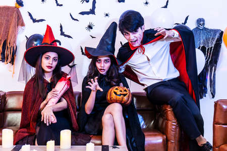 Asian Happy friends sitting on sofa in costumes and makeup on a celebration of Halloween posing with...