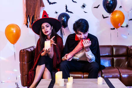 Asian Happy friends sitting on sofa in costumes and makeup on a celebration of Halloween posing Eat fake human legs in party, Group of young people enjoying Halloween party with many items at home or nightclub.の写真素材