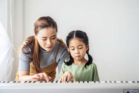 Happy little Asian deaughter playing piano with mother at home, Mother teaching daughter to play...