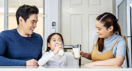 Happy family having fun in the kitchen. Asian Father, mother and little daughter spending time together and having breakfast drinking and hold glasses of milk at tableの写真素材