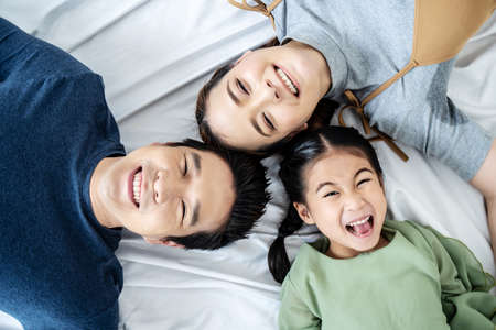 Top view of Happy Asian beautiful young mother, father and their daughter looking at camera and smiling while lying on bed head to head in bedroomの写真素材