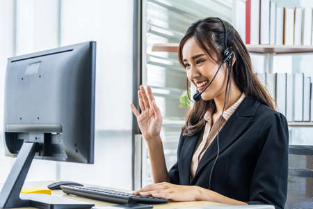 Smiling friendly asian businesswoman with headset working and greets interlocutor for online communication. Remote learning worldwide conceptの写真素材