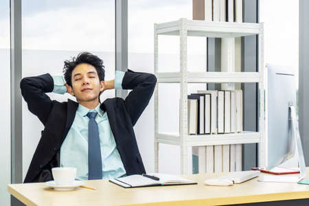 Young asian businessman having break and resting holding hands behind head and keeping eyes closed while sitting at his working placeの写真素材