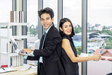 Businessman and businesswoman stand back to each other in suits smiling at the camera and working on laptops in officeの写真素材