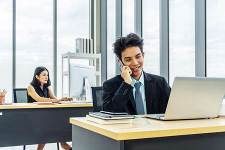 Young businessman sitting at office talking on mobile phone, sitting at desk, using laptop, smiling, businesswoman working on laptop in her workstation in background.の写真素材