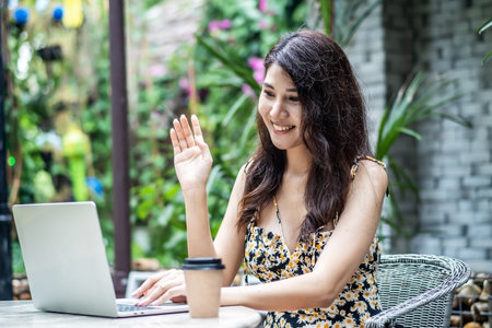 Beautiful asian young woman making online video call via laptop application. She says hi and waves her hand towards the camera at the cafe outdoorの写真素材