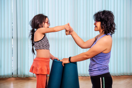 Asian woman and cute little girl are looking at each other and laughing while holds yoga mat after exercise in gymの写真素材
