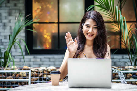 Beautiful asian young woman making online video call via laptop application. She says hi and waves her hand towards the camera at the cafe outdoorの写真素材