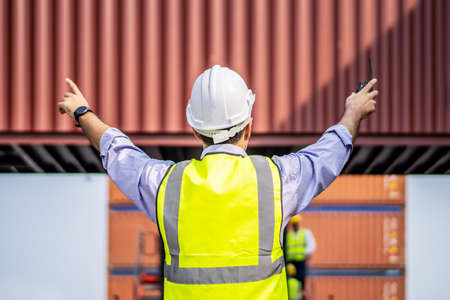 Rear view of engineer in reflective clothing with safety helmet is working and checking at containers cargo on shipping port area at harborの写真素材