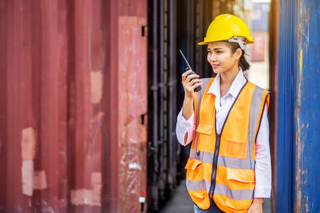 Young confident woman engineer smiling and using radio communication and wearing yellow safety helmet and check for control loading containers box from Cargo freight ship for import and exportの写真素材