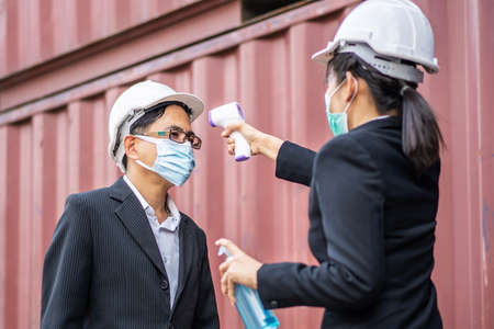Female foreman wearing a mask measures the temperature for a male foreman in a suit. They stood and waited for the measurement with a non-contact infrared thermometer to prevent covid virusの写真素材