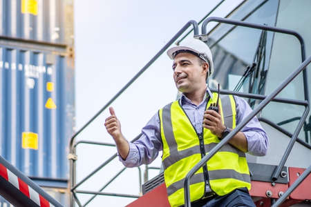 Foreman in hard hat show thumb up signal and using walkie talkie control loading Containers box from Cargo freight ship in Warehouse shipping transportation conceptの写真素材
