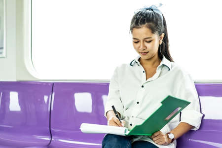 Beautiful asian woman working urgently writing on sheet of paper in a clipboard with pen at the electric train in the morningの写真素材