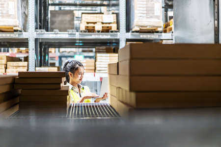 Asian female worker in safety vest sitting and working with computer laptop in storage warehouse. people, warehouse and industry conceptの写真素材