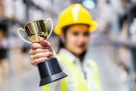 Happy woman warehouse worker holding a trophy after being selected as an outstanding employee, Selective focus of hand hold trophyの写真素材