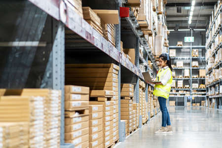 Asian engineer woman check stock details on tablet computer for checking boxes with logistics on shelves with goods background in warehouse, logistic and business exportの写真素材
