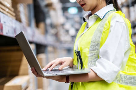 Asian engineer woman check stock details on tablet computer for checking boxes with logistics on shelves with goods background in warehouse, logistic and business exportの写真素材
