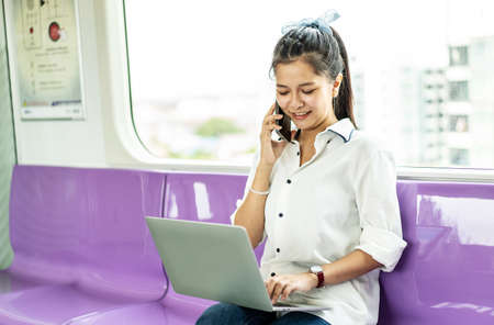 Asian businesswoman talking on cellphone and working on laptop while traveling by sky train. Business travel concept.の写真素材