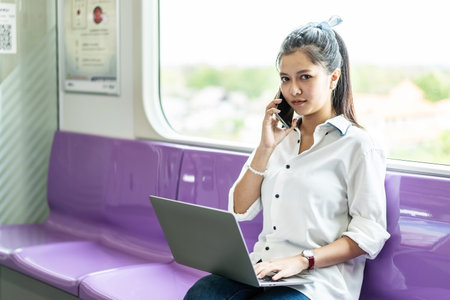 Asian businesswoman talking on cellphone and working on laptop while traveling by sky train. Business travel concept.の写真素材